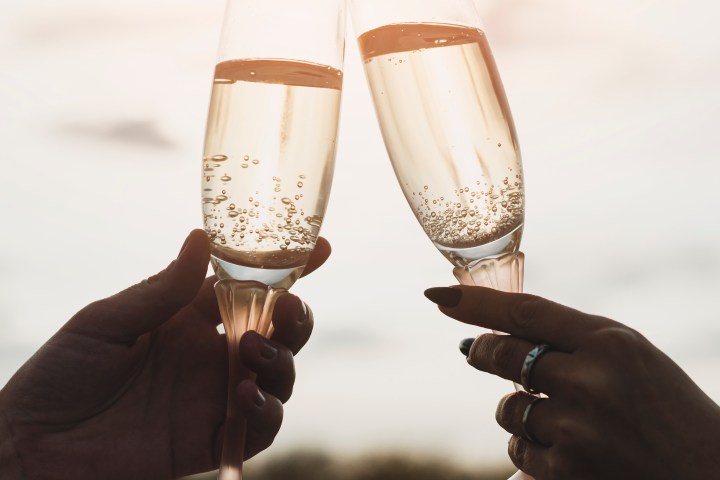 Two hands clinking champagne glasses against a sunset sky background.