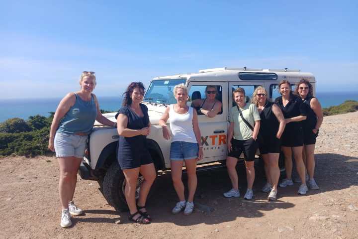 Eight women posing by a white off-road vehicle near the coast.