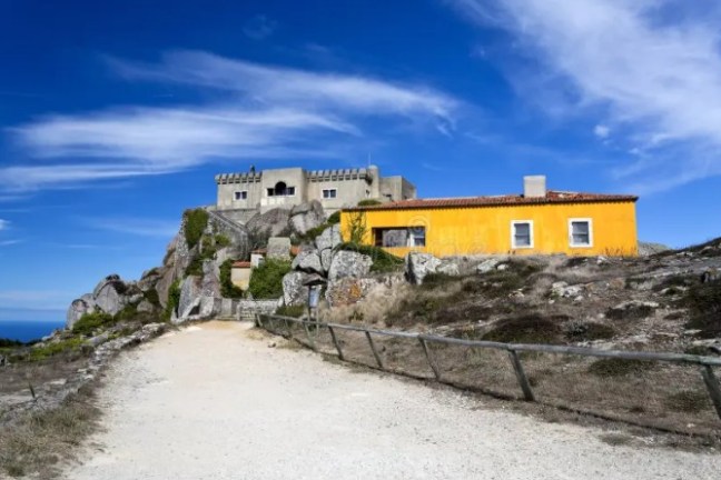 Path leading to stone buildings on a rocky hill under a blue sky.