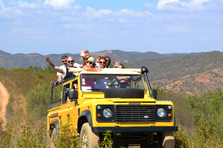 Group of people in a yellow jeep on a dirt road with mountains in the background.