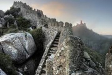Ancient stone fortress with crenelated walls and stairs, overlooking a misty landscape with a distant castle.