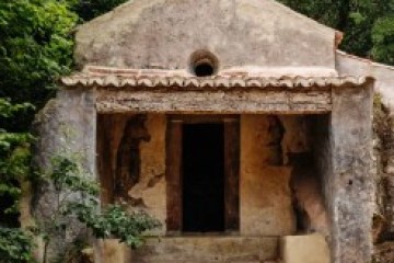 Small stone chapel with a cross on top, nestled among lush green trees, with worn steps leading to the entrance.