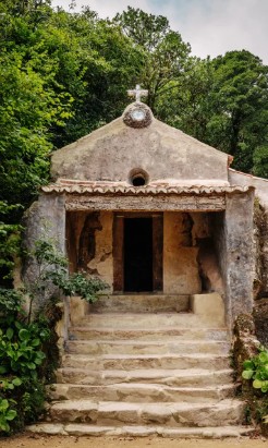 Small stone chapel with a cross on top, nestled among lush green trees, with worn steps leading to the entrance.
