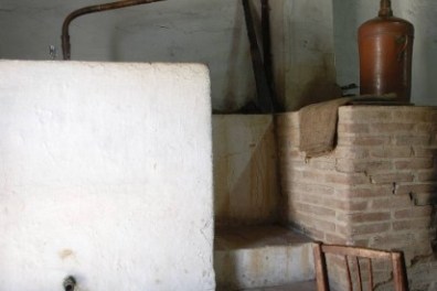 Rustic distillation setup with copper pipes and a wooden chair in a stone-walled room.