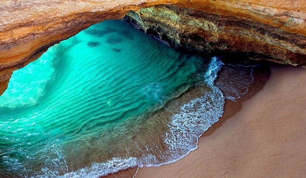 Sea cave with turquoise water and sandy shore illuminated by sunlight.