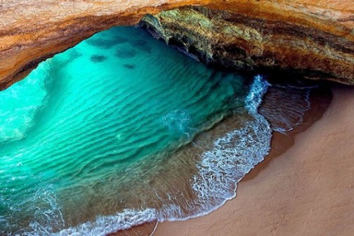 Sea cave with turquoise water and sandy shore illuminated by sunlight.