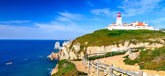 Lighthouse on a cliff by the sea under a blue sky with a wooden fence path.