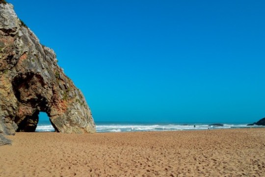 Beach with rock arch, sandy shore, and clear blue sky.