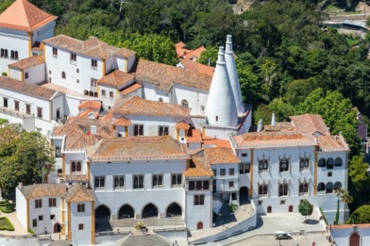 Aerial view of a castle with red-tiled roofs and two large conical chimneys in a forested area.