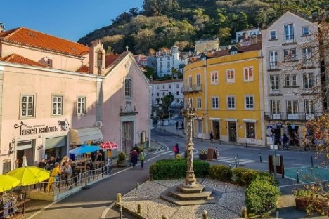 Colorful European square with cafes, vibrant buildings, and a hill backdrop on a sunny day.
