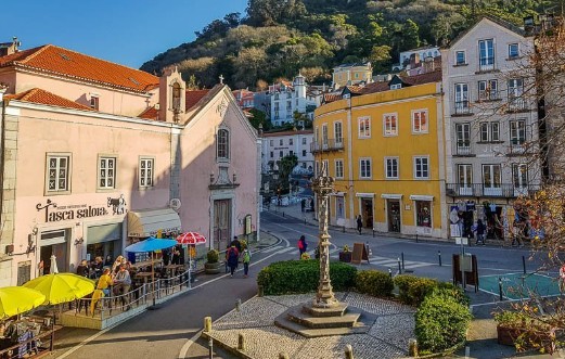 Colorful European square with cafes, vibrant buildings, and a hill backdrop on a sunny day.