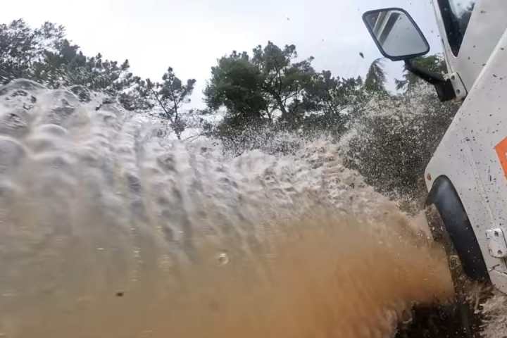 Truck driving through deep mud, water splashing dramatically by forested area.