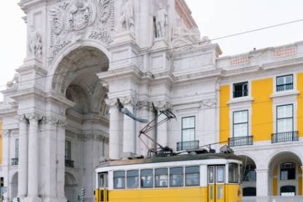 Yellow tram passing in front of an ornate stone arch, with yellow buildings in the background.
