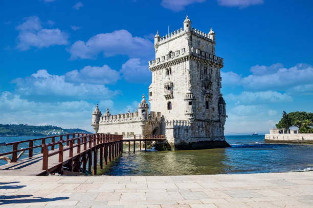 Historic stone tower beside water, with a wooden walkway leading to it under a blue sky with clouds.