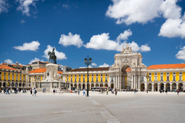 City square with statue, arch, yellow buildings, and people under a blue sky with clouds.